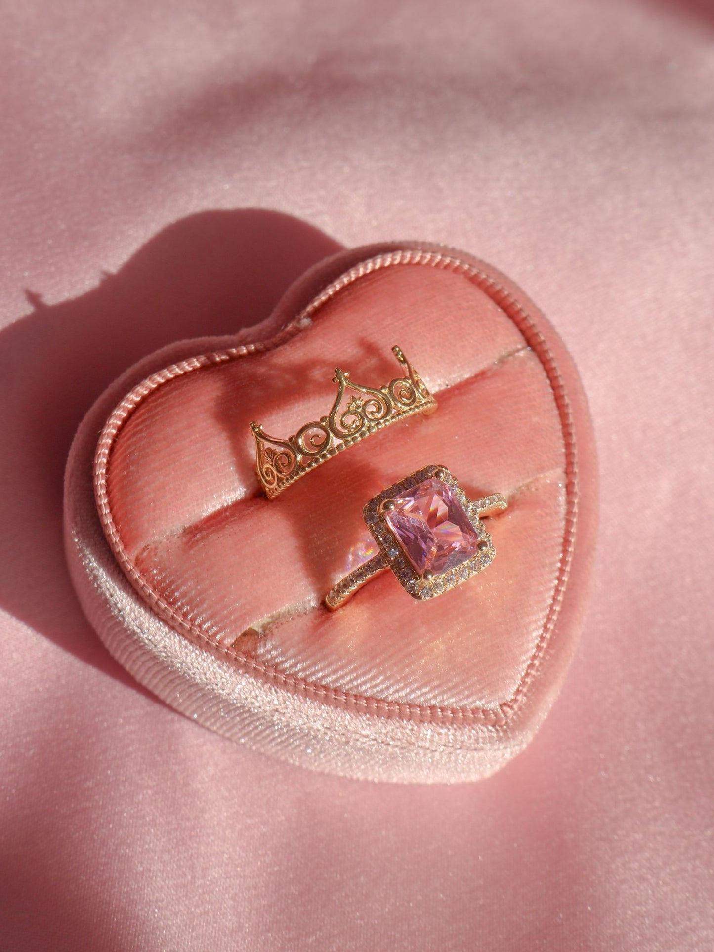 Two rings, one with a large pink gemstone and the other with a smaller clear stone, placed on a pink cushion with a heart shape.