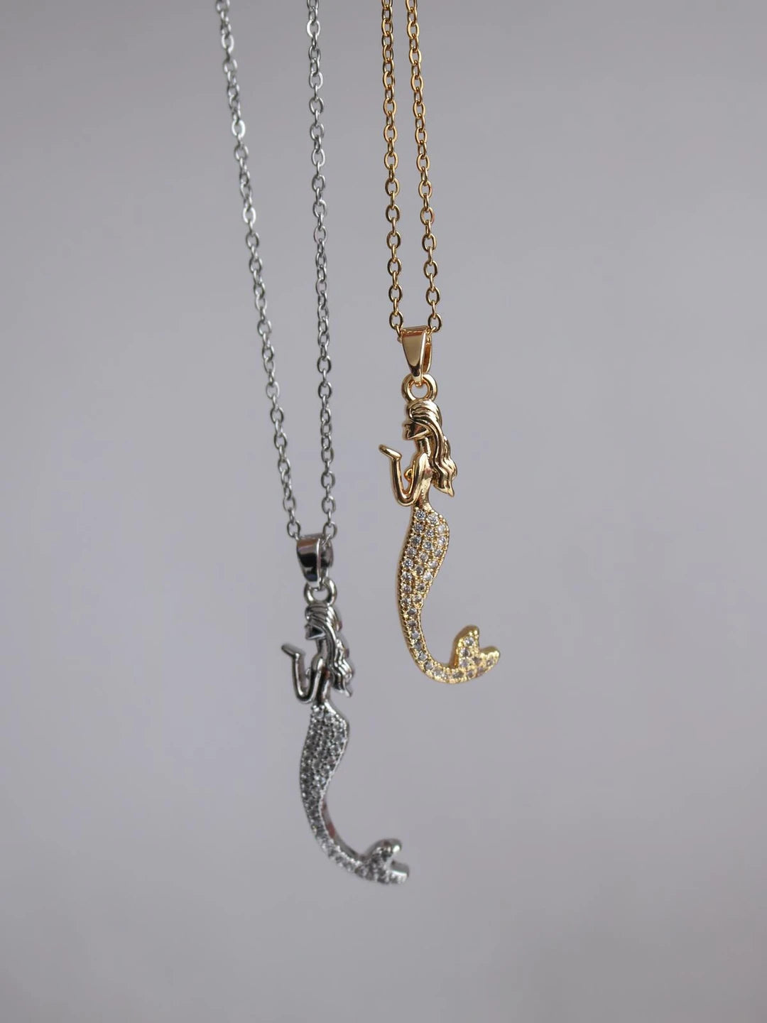 Two mermaid-themed necklaces, one with a silver mermaid figure attached to a chain and another with a gold mermaid figure attached to a chain.