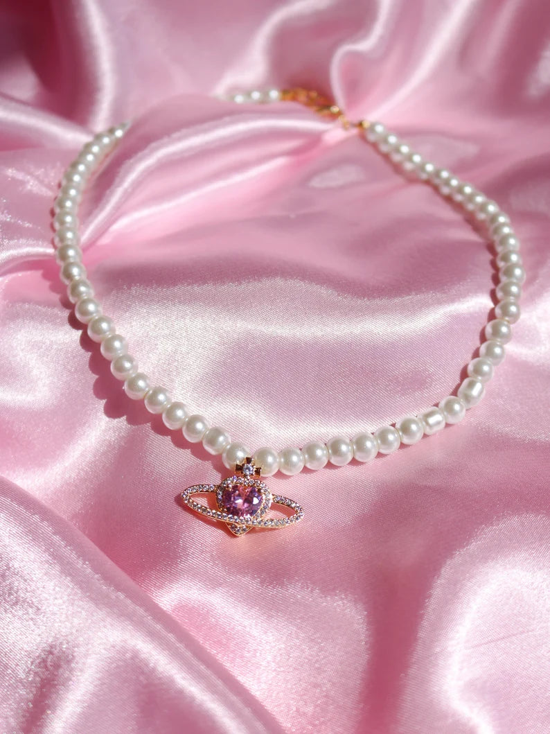A gold-plated necklace with a heart-shaped saturn pendant and pearl-like beads displayed on a pink satin surface.