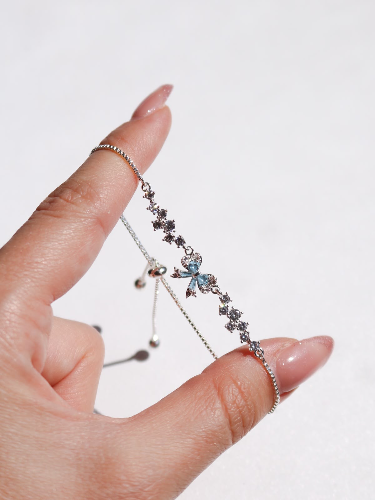 A delicate silver-toned chain bracelet with a small blue bow charm and dark star-like accents, held on a person's finger against a plain light background.