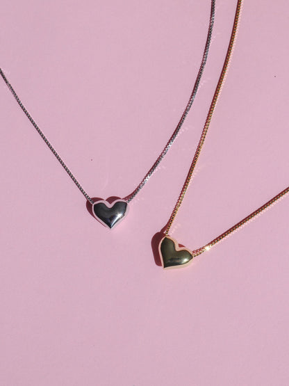 Two necklaces with heart-shaped pendants, one silver and one gold, displayed against a pink background.