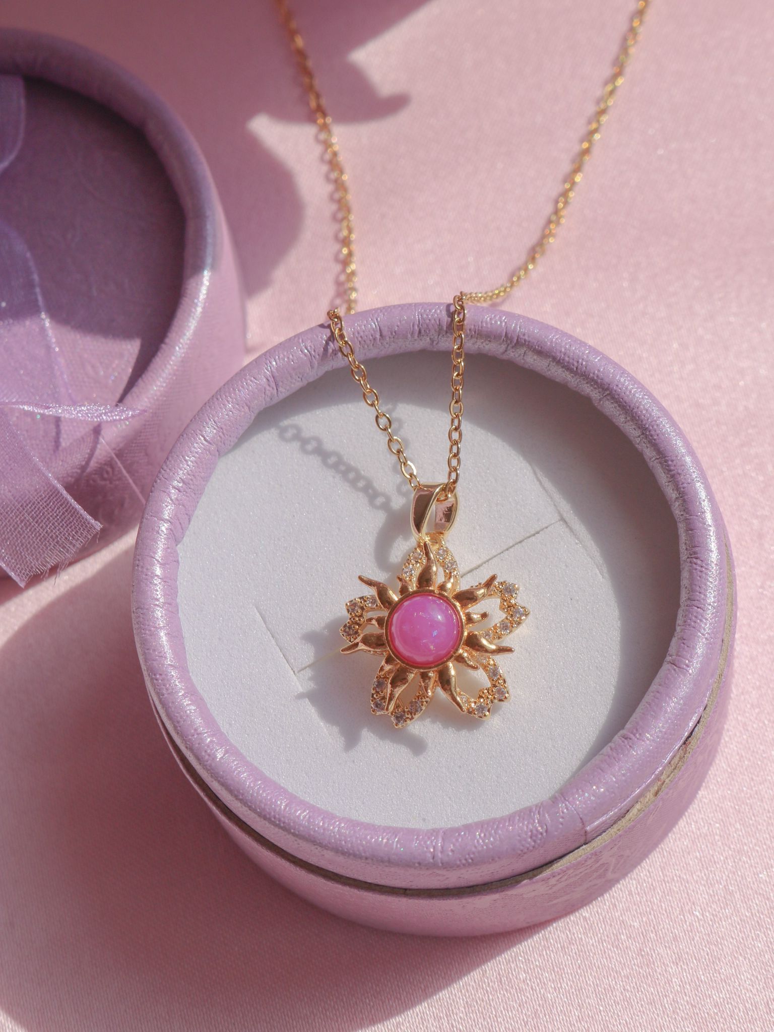 A gold-toned spinning necklace with a pink and white sundrop flower design, presented in a purple jewelry box.