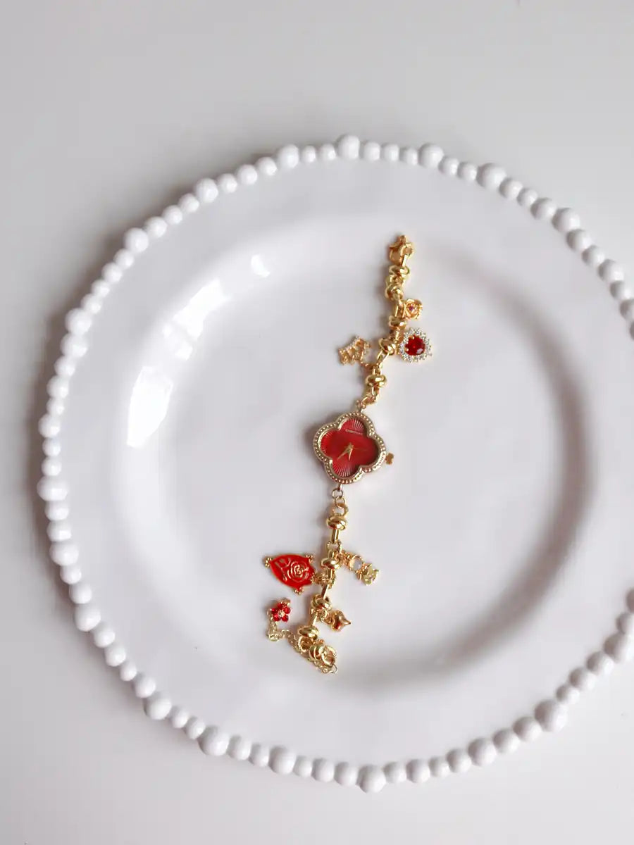 Belle Charm Watch  gold chain bracelet with red heart and leaf charms, displayed on a white beaded ceramic plate against a light gray background.