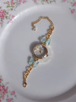 A gold-tone chain watch bracelet with a round dial, decorated with light blue crystals and bow charms, resting on a white plate with a pink floral border.