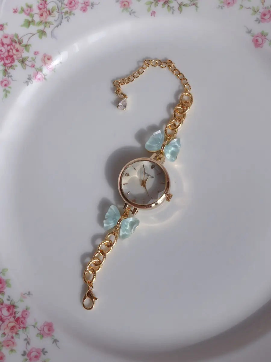 A gold-tone chain watch bracelet with a round dial, decorated with light blue crystals and bow charms, resting on a white plate with a pink floral border.