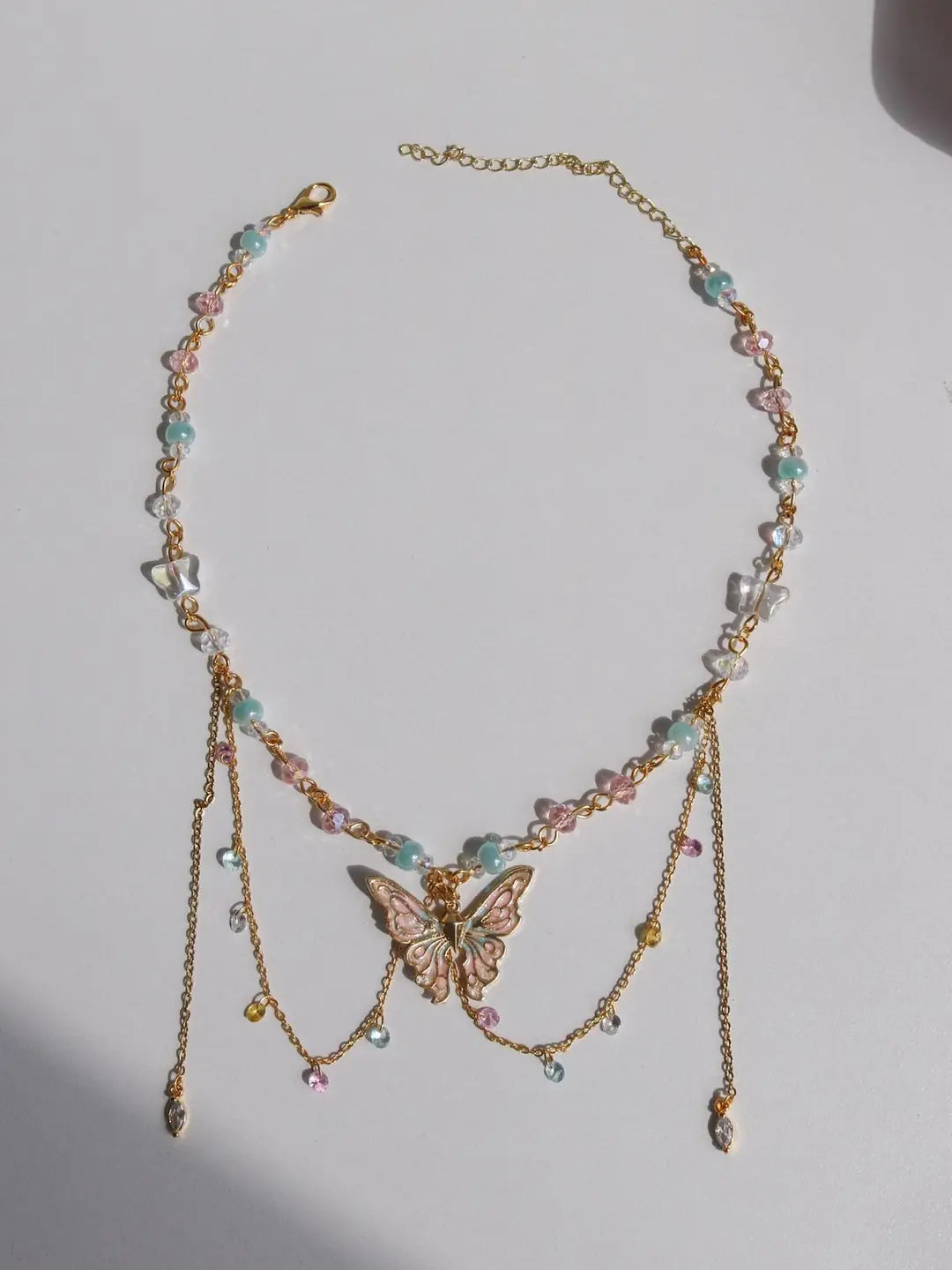 A gold-toned beaded necklace with a central butterfly charm, featuring pastel pink, mint green, and white beads, displayed on a light gray surface.