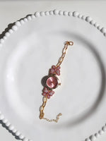 A gold-tone chain bracelet watch with a round pink dial and two pink butterfly charms, displayed on a white beaded plate.