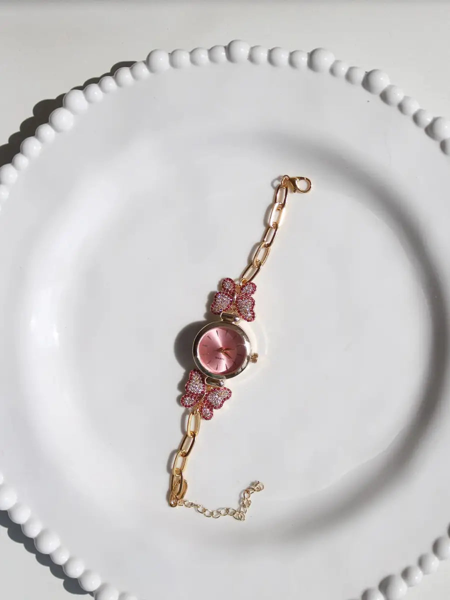 A gold-tone chain bracelet watch with a round pink dial and two pink butterfly charms, displayed on a white beaded plate.