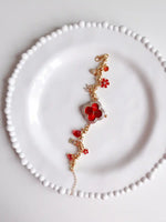 A delicate gold chain bracelet with red crystal elements, including a central four-petal flower and small floral accents, displayed on a white beaded plate.