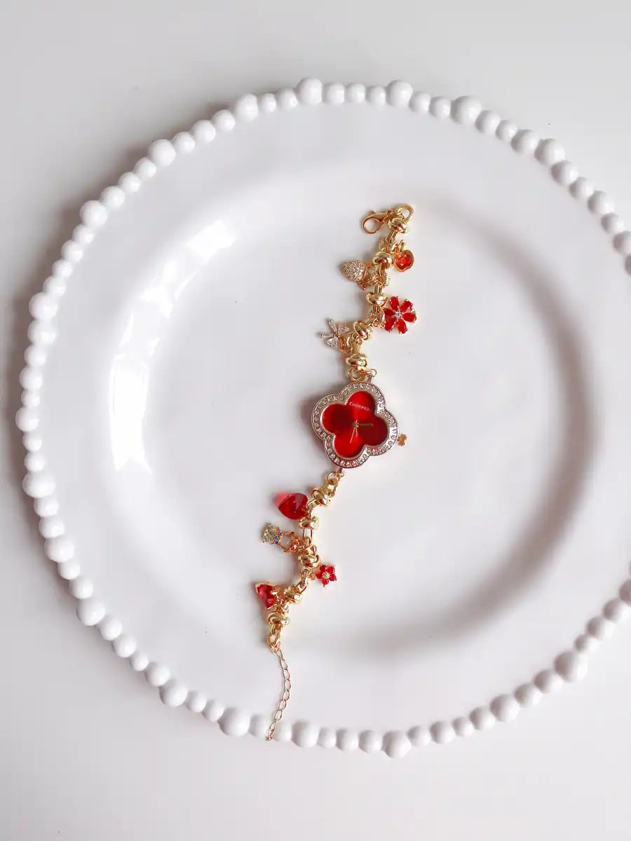 A delicate gold chain bracelet with red crystal elements, including a central four-petal flower and small floral accents, displayed on a white beaded plate.