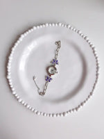 A silver-toned chain bracelet with purple flower charms and a round clock face, displayed on a white plate with a beaded rim.