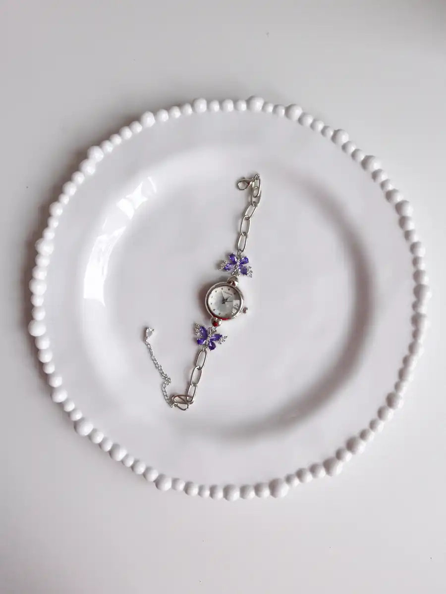 A silver-toned chain bracelet with purple flower charms and a round clock face, displayed on a white plate with a beaded rim.