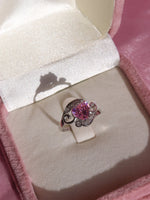 An engagement ring with a silver band and a large pink central stone, possibly a zirconia, with silver accents around it, displayed in a pink box.