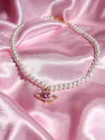 A gold-plated necklace with a heart-shaped saturn pendant and pearl-like beads displayed on a pink satin surface.